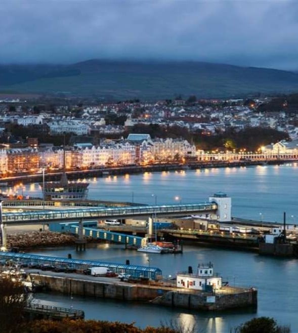 Evening view of Douglas Harbour on the Isle of Man, showing illuminated waterfront buildings, calm water, moored ferries and boats, with rolling hills in the background.